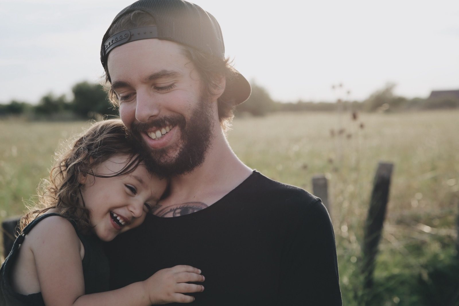 A father laughing with his young daughter, her head resting on his shoulder, in a sunlit field